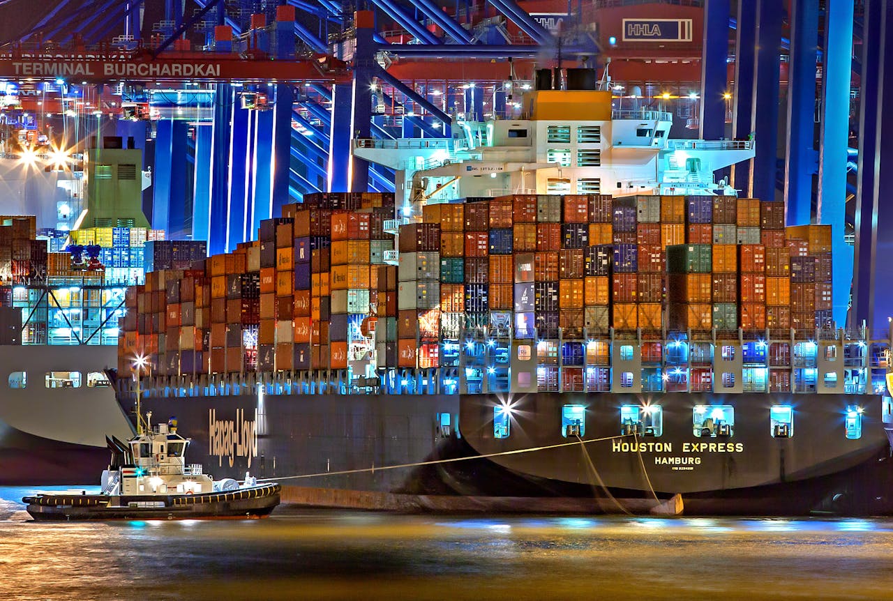 Services A brightly lit cargo ship at Hamburg harbor with stacked containers and a tugboat.